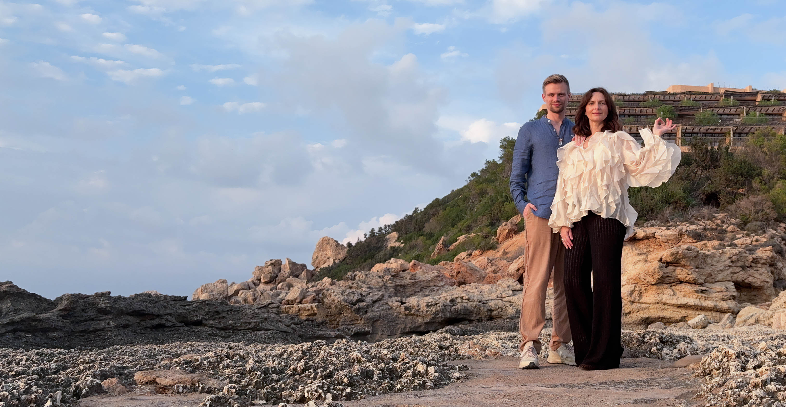 Man and woman standing together on rocky terrain with hills and partly cloudy sky in the background.